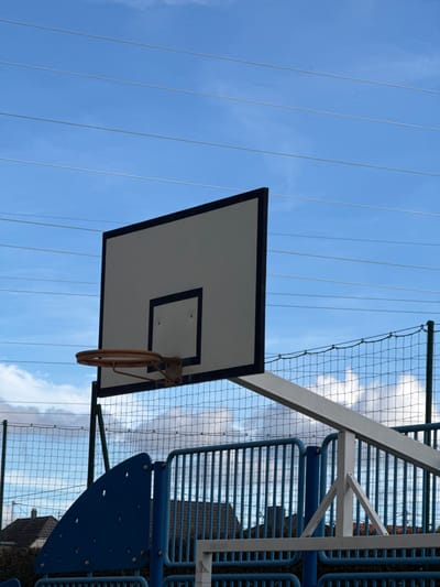 The image displays an outdoor basketball setup located in Niederhausbergen, France, under a partially cloudy blue sky. A white rectangular basketball backboard, outlined in black, dominates the center, featuring a smaller black-bordered rectangle directly below its center. An orange-brown metallic basketball rim is attached to the backboard. The backboard is supported by a dark green metal post on the left and a white angular metal support structure on the right. Behind the basketball apparatus, a fence with black netting and green posts is visible. Below the backboard, a blue playground structure featuring a curved panel on the left and vertical metal bars on the right is present. Multiple thin, light-colored electrical wires traverse horizontally across the upper sky. In the background, through the fencing and structures, parts of dark-roofed residential buildings and dark green foliage are discernible. No individuals are present in the scene.