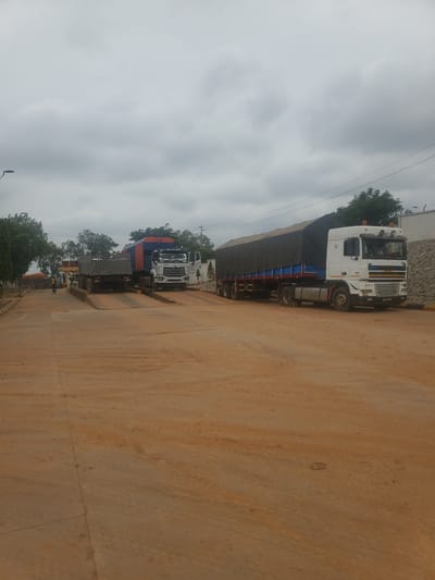 In Gwagwada, Nigeria, on a cloudy day, two large trucks are positioned on truck scales on a dirt road. The truck on the left is a dump truck with a blue and orange container. The truck on the right is a semi-trailer truck with a black tarp covering its cargo. The truck scales are elevated, with ramps leading up to them. A person wearing a high-visibility vest is standing near the truck on the left, possibly overseeing the weighing process. The environment appears to be an industrial or transportation hub, with trees and buildings visible in the background. The ground is covered in reddish-brown dirt, suggesting it may be a dry or arid region.