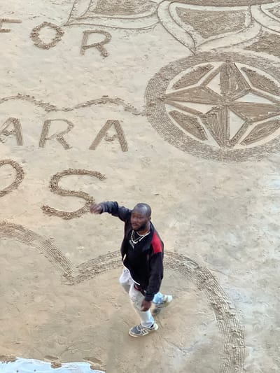 An aerial, high-angle view shows a man in casual clothing on a sandy beach in San Sebastián, Spain. He is wearing a black jacket with red accents on the sleeves, light-colored pants, and athletic shoes. The man is walking and gesturing with his right hand. In front of him, etched into the sand, are large letters that appear to spell "FOR AMARA OS" and decorative patterns, including a compass rose and a heart shape. The beach is wet and the sand shows textures from the etching process.