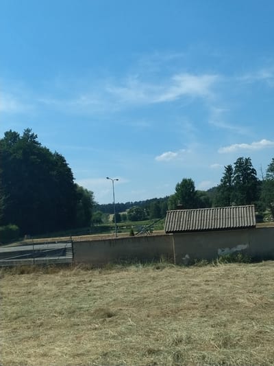 The image shows a rural scene under a clear blue sky with sparse white clouds. In the foreground, there is a field of dry, yellowish grass. A low, rough concrete wall runs horizontally across the middle ground, topped with a corrugated metal roof structure. Behind the wall, there is a fenced area with a paved path and what appears to be steps or a ramp. Further back, a dark green, dense forest covers a hill. A single street lamp with two lights stands to the left of center. In the distance, rolling hills and more trees are visible, with a body of water, possibly a pond or lake, to the right of the street lamp. The overall impression is a serene, somewhat arid landscape on a sunny day, likely in the countryside near Horažďovice, Czechia.