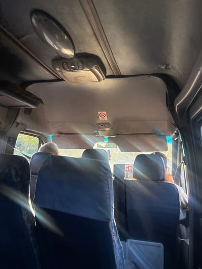 The image depicts the interior of a vehicle, likely a minibus or shuttle, in Partskhanakanevi, Georgia, viewed from a rear passenger seat looking towards the front. The light gray headliner includes an oval chrome-finished light fixture and a beige plastic overhead control unit with red indicator lights. Two beige sun visors are visible, one positioned centrally and another angled to the right. A red and white circular "No Smoking" sign, displaying a crossed-out cigarette, is affixed to the headliner above the right sun visor. The backs of several dark blue passenger seats are prominent in the foreground and midground. Sunlight intensely streams through the front windshield, creating significant glare and lens flares across the scene. The driver, identifiable by the back of a head with short gray hair, is visible in the front left seat. A second individual, with the back of their head and shoulder visible, occupies the front right passenger seat, partially obscured by the glare, with a hint of an orange garment. Through the windows, green foliage is discernible outside the vehicle.