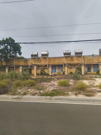 A long, one-story building with a yellow, weathered facade stretches across the frame, viewed from a slight side angle. The building has multiple windows, some with blue panes and others with barred sections. A low, yellow-capped fence runs in front of the building. Several large, cylindrical, silver water tanks are mounted on metal frames atop the roof of the building. To the left of the building, a large tree with dense green foliage is visible. The foreground is dominated by a grey road with a white stripe on its left edge, suggesting the photo was taken from a vehicle moving along the road. The ground in front of the building is covered in dry, sandy earth with sparse patches of dry grass and shrubbery. The sky above is overcast and pale grey, with several dark, horizontal power lines crossing the upper portion of the image. The scene appears to be in a rural or semi-urban area, possibly a school or administrative building, given the infrastructure. The location is identified as Bac Cam Ranh Ward, Vietnam.