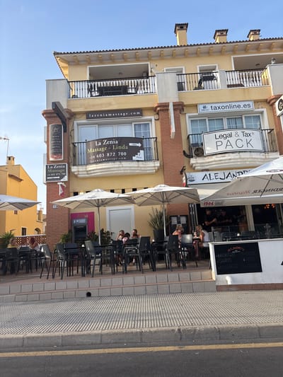 The image shows a street-level view of a yellow building with balconies and awnings in Urbanización La Cenia II, Spain. The building houses several businesses, including a massage salon with signage for "La Zenia Massage - Linda" and a website address "lazzeniamassage.com". Next to it is a sign for "Legal & Tax PACK" and "taxonline.es". A restaurant named "ALEJANDRO'S BAR RESTAURANTE" is visible at street level with outdoor seating. Several people are seated at tables outside the restaurant under large white umbrellas, some appearing to be dining or socializing. An ATM machine is also visible. The building has a light blue sky above and a sidewalk and street in the foreground.