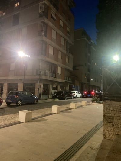 The image depicts a street scene at dusk or night in the city of Ancona, Italy. A multi-story building with residential or commercial units dominates the left side of the frame. Its facade features rows of windows, some lit from within, and balconies. A street light illuminates the sidewalk and a portion of the street. Several cars are parked along the street, including a blue hatchback on the left and darker sedans and SUVs further down. A white car with a "P" parking sign above it is also visible. The sidewalk is paved with stone and concrete blocks, and there are several cube-shaped bollards. In the background, another building is visible, and trees line the right side of the frame, with foliage partially obscuring the view. The sky is a deep blue, indicating twilight or early night. The overall atmosphere is quiet and urban.