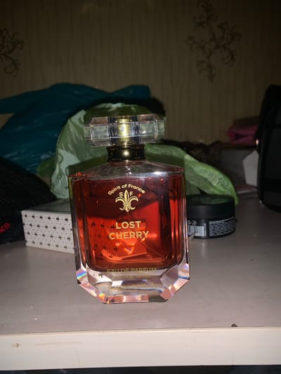 A bottle of "Lost Cherry" perfume is the primary subject of the image, positioned on a shelf in what appears to be a room. The bottle is made of glass, with a red liquid inside and a faceted crystal cap. The label on the bottle reads "Spirit of France S F" and "LOST CHERRY EAU DE PARFUM NATURAL SPRAY."
Behind the perfume bottle, several items are visible, including what seems to be a green plastic bag, a white box with a pattern of dark crosses, and a black jar with a label. The background is a wall with a textured wallpaper and a decorative pattern. The lighting suggests an indoor setting, likely in the evening or in a dimly lit room. The overall scene is a still life arrangement of personal items.