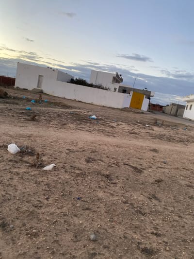 The image displays an outdoor scene in الغناذة, Tunisia, primarily featuring a large expanse of unpaved, sandy-dirt ground. This ground appears dry and uneven, scattered with small rocks, pebbles, and sparse, dry patches of vegetation. Numerous pieces of litter, notably white and blue plastic bags or fragments, are visible across the dirt, suggesting an unkempt environment.
To the left, a cluster of low-rise buildings with flat roofs is visible. These structures are predominantly white, with one prominent building featuring a distinct yellow section on its upper facade. The architecture suggests residential or simple utility buildings common to the region.
The sky occupies the upper left portion of the frame, showing a mix of pale blue and soft, elongated clouds, indicative of either early morning or late afternoon light. The weather appears partly cloudy with diffused illumination. No people or specific activities are depicted. There is no visible text. The overall scene portrays a quiet, semi-developed or undeveloped area characterized by its arid terrain and simple buildings, with noticeable environmental litter.