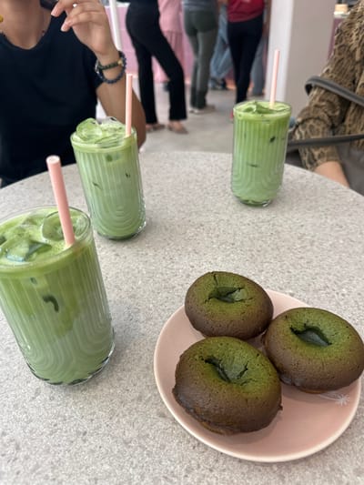 The image displays an indoor scene within a cafe or dessert establishment located in Vandœuvres, Switzerland. The foreground is dominated by a light grey, speckled table surface. Arranged on the table are three clear glass tumblers, each filled with a vibrant green, iced beverage, presumably matcha, and containing a pink straw. One tumbler is positioned in the immediate foreground, another to its right, and a third further back on the right.
To the right of the tumblers, a small, light pink ceramic plate holds three circular, dark green baked goods, appearing to be matcha-flavored donuts or financiers, each with a darker brown exterior and a distinct central indentation.
On the left side of the frame, the upper torso and left arm of an individual wearing a black short-sleeved top are visible. A blue and gold beaded bracelet adorns their left wrist, and their hand is raised near their face. On the right, a portion of another individual's arm and shoulder is visible, clad in a brown and black patterned garment, with a black strap indicative of a bag. The background shows blurred figures of multiple individuals standing, suggesting a busy public environment.