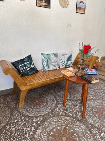 The image depicts a static seating area, possibly an interior or semi-outdoor space, in Panglao, Philippines. A light-brown polished wooden slatted bench, featuring a curved left armrest and a raised right backrest, is positioned against a white textured wall. Four decorative throw pillows are arranged on the bench: one dark green with "Tropical Dream" text and palm leaves, and three white with green palm leaf patterns and various phrases. A small, round, dark brown wooden side table with three legs stands in front of the right section of the bench. Adorning the table are a woven brown and tan vase holding artificial red flowers and green foliage, along with several books, including visible titles such as "November 9" and one with an orange cover. The white wall above the bench displays three framed pictures—one of a water splash, another of a jumping dolphin, and a third ambiguous image—alongside a light-colored, irregularly shaped decorative object and a white electrical outlet. The floor is tiled with a repeating circular mosaic pattern in shades of brown, white, and grey. No individuals, actions, or interactions are present in the scene.