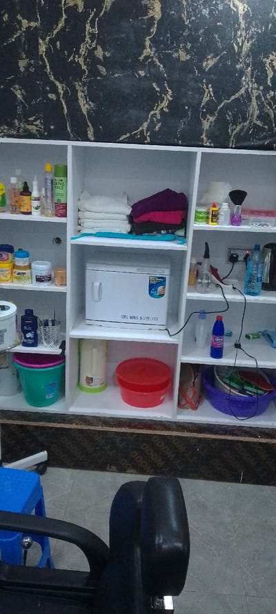 The image displays an interior view of what appears to be a beauty salon or barbershop located in Matathani ward, Kenya. The scene is dominated by a white modular shelving unit, meticulously organized with various beauty and hair care products. The wall behind the shelving unit features a black surface with golden, marble-like veining. The floor is composed of grey tiles, and a dark brown baseboard with "WOOD TY" text runs along the bottom.

On the top shelf, from left to right, various clear and colored bottles containing liquids, possibly shampoos, conditioners, or oils, are arranged. Next to them are stacks of neatly folded white towels and a separate stack of colored towels (purple, pink, dark red). A long, turquoise object lies on the shelf. The rightmost section of the top shelf contains smaller items including a white container, small bottles, a purple-handled brush, and a clear plastic container with small accessories.

The middle shelf features assorted jars and tubs of creams or balms, including a "BABYCARE" product, on the left. Centrally, a white "TOWEL WARMER UV STERILIZER" appliance is visible, with a blue label indicating "For Hair Salon & Nail Shop." A black power cable is plugged into an outlet on the right side of this shelf, connecting to the sterilizer and other visible devices. A clear spray bottle, a red and white power adapter, and a blue plastic water bottle are also present on the right section of the middle shelf.

The bottom shelf on the left holds a white cylindrical container, a blue "NIVEA" bottle, a metallic holder with dark tools, and a green plastic bucket with a pink rim. The middle section of the bottom shelf contains a white cylindrical container with a green base and a red plastic tub with a lid. The rightmost section of the bottom shelf features a blue plastic basin containing miscellaneous items, a blue bottle labeled "TOPEX," and a light blue cloth.

In the foreground, a portion of a black salon chair with a chrome lever is visible, along with a blue plastic stool. No individuals are present in the image.