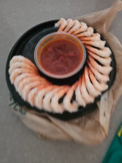 A close-up, high-angle shot shows a black plastic platter filled with peeled, cooked shrimp arranged in a semi-circle around a clear plastic container of red cocktail sauce. The shrimp are pinkish-white and appear to be chilled. The platter is resting on a brown paper bag, which is on a gray, textured surface, likely concrete. The background is out of focus, but suggests an outdoor setting. The image is blurry and has a shallow depth of field. There are no people visible in the image. The scene is consistent with a casual outdoor meal or snack in Islamorada, United States of America.