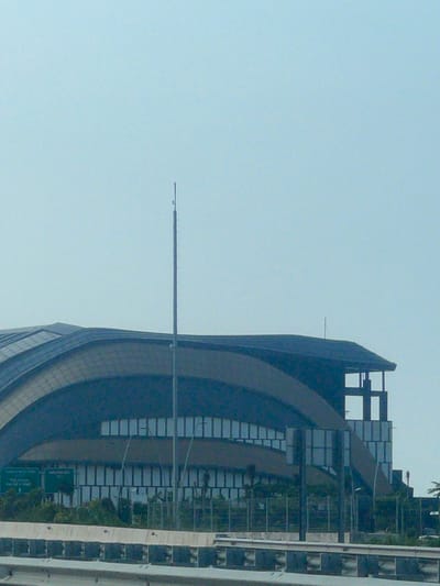 The image captures a segment of a modern, curved building with a dark, reflective facade, possibly an airport terminal or a large convention center. The building features numerous rectangular windows arranged in rows, with some sections of the facade appearing to be made of tinted glass. A prominent, slender antenna or mast stands vertically in front of the building. In the foreground, a concrete barrier with a reflective surface, likely part of a highway or overpass, is visible. Lush green foliage and trees are present behind the barrier and in front of the building. Several green signage boards are mounted on poles in front of the building. The sky is a pale, uniform blue, suggesting a clear day. The overall scene appears to be a transportation hub or a large public facility in an urban or suburban setting. The building's architecture suggests a contemporary design, likely built in the late 20th or early 21st century. The presence of a highway barrier and signage further indicates a location with significant traffic flow and public access.