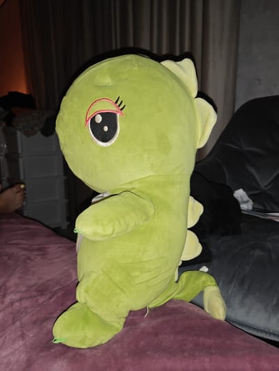 A medium shot shows a bright green plush toy dinosaur sitting on a deep plum-colored blanket. The dinosaur is facing left, with its head turned slightly towards the camera. It has a large head with a single prominent eye on the left side. The eye is black with a white pupil and outlined in white and pink, with three black eyelashes. The dinosaur's body is plump, and it has small, stubby arms and legs. Yellow-colored spines run along its back. In the background, the scene is dimly lit. To the left, there is a white plastic chest of drawers and a dark curtain. To the right, part of a dark grey cushioned sofa is visible. A bare foot with yellow toenails is visible on the far left of the image, resting on the plum blanket. The overall environment suggests an indoor setting, possibly a bedroom or living room, in Izmail, Ukraine.