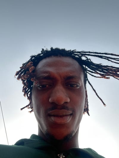 An individual with dark skin and dreadlocks is captured in a low-angle, close-up portrait in Ilishan, Nigeria. The subject, appearing male, looks directly into the camera with a neutral expression. Dreadlocks are visible on the head; some on the left side are shorter and clustered, while those on the right are longer and appear to be swept towards the right, suggesting environmental airflow. Minimal stubble is present on the chin and upper lip. The subject wears a dark green hooded garment, revealing a silver-colored zipper pull at the collar. The background is a bright, largely clear sky, transitioning from a lighter glow on the lower left to a faint blue-grey towards the top. A thin dark line, possibly a wire, is visible vertically along the far left edge of the frame.