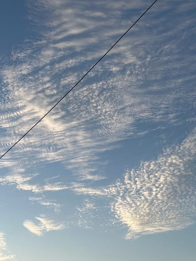 This is a photograph of a bright blue sky filled with wispy white clouds. A thin, dark wire stretches diagonally across the frame from the upper left to the lower right. The clouds appear to be altocumulus formations, characterized by their patchy, rounded appearance. The lighting suggests it is daytime, possibly late afternoon due to the warm tones in some of the clouds. There are no people, buildings, or any other identifiable structures or objects visible in the image, besides the wire, which is likely a power line or telephone wire. The location is specified as Slobozia, Moldova, however, the image itself provides no contextual visual cues to confirm this specific location; it is a generic sky scene.