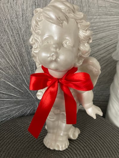 This is a close-up, eye-level, medium shot of a white cherub figurine with curly hair, plump cheeks, and closed eyes. A bright red satin ribbon is tied in a bow around its neck, with one end hanging down. The figurine is standing on a dark gray textured surface, possibly a placemat or cushion. Behind the figurine, a shimmering, silvery fan-shaped decoration with a textured, grooved surface is visible. The lighting appears to be soft, highlighting the pearlescent sheen of the figurine and the glossiness of the ribbon. The background is out of focus, suggesting a shallow depth of field. The scene is static and appears to be an indoor setting. There is no indication of the location in Balabanovo, Russia.