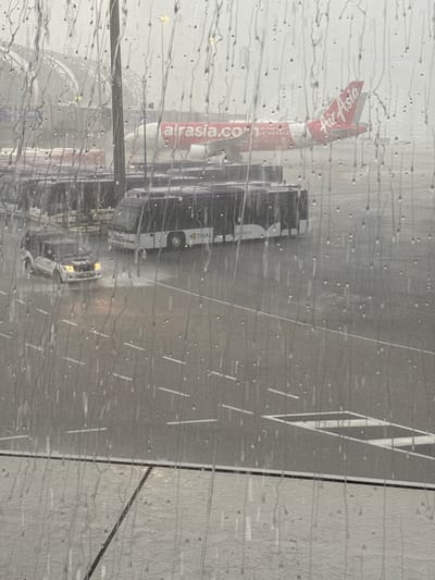 This image shows an airplane viewed through a window covered in raindrops. The airplane is red and white, with "AirAsia.com" printed on its side. Behind the AirAsia plane, several white buses with "THAI" written on them are parked. The scene appears to be an airport tarmac in Nong Prue Subdistrict, Thailand. The weather is rainy, indicated by the water droplets on the window and the wet appearance of the tarmac. The lighting suggests it is daytime, possibly overcast due to the rain. The overall mood is somewhat subdued, likely due to the weather.
