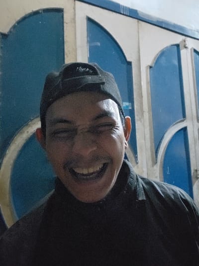 A person with a dark complexion, wearing a black cap and a dark top, smiles broadly with their eyes closed. The background features a blue and white structure with curved shapes. The image is taken from a low angle and is slightly rotated. Light reflects off the person's forehead, emphasizing their smile and the curve of their nose.