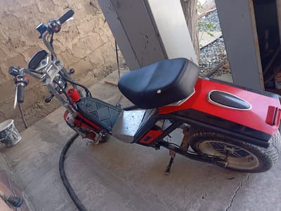 The image shows a red and black electric scooter parked on a concrete surface in a yard. The scooter is upright and appears to be in working condition. The scooter has a black seat, a digital display on the handlebars, and a metal platform where the rider would place their feet. There is a bucket to the left of the scooter and a black hose on the ground in front of it. In the background, there is a wall made of rough stones and a large metal structure. A tree trunk is visible behind the metal structure. There is a pile of gravel and some vegetation on the ground in the background. A can of what appears to be a cleaning product is visible to the right of the frame, near the metal structure. The overall environment suggests an outdoor storage or maintenance area. The location is specified as the city of Ararat, Armenia.
