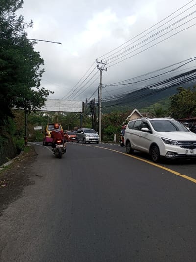 The image depicts a street scene in Saing Ta Mawt Lay, Myanmar, under an overcast sky. A two-lane asphalt road with a solid yellow line on the right curves slightly to the left. In the foreground, a person wearing a red long-sleeved shirt and a light-colored helmet rides a dark motorcycle away from the camera. Behind the motorcycle and to its right, a white SUV with license plate "F 1427" is also moving away from the camera. Further down the road, an orange minivan is partially visible on the left, and a silver minivan and an orange car are approaching the camera in the opposite lane. Utility poles with numerous overhead power lines are prominent on the right side of the road. A white, rectangular overhead sign structure spans the road in the mid-ground. The left side of the road features a concrete-lined drainage ditch and a vegetated embankment. In the background, dark, tree-covered mountains rise under the cloudy sky, with several houses featuring light-colored roofs nestled among the trees.