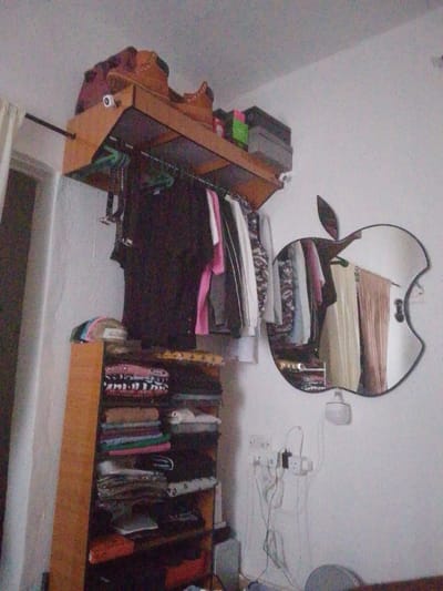 The image displays an interior room in Eziobodo, Nigeria, featuring white painted walls. A light-colored wooden, wall-mounted storage unit occupies the upper left and central portions of the frame, extending horizontally. This unit includes an upper shelf holding decorative wooden items and three stacked boxes, one green and two grey. Below this shelf, a black metal rod functions as a clothes rail, supporting various garments on plastic hangers, including dark shirts, pink clothing, and patterned items, along with several black belts.
To the left and beneath the wall-mounted unit, a tall, narrow, light-colored wooden shelving unit is visible. It holds multiple stacks of neatly folded clothing in various colors and patterns, topped by a beige cap with visible branding.
On the right wall, an Apple-shaped mirror is mounted, reflecting a section of the room that includes light-colored curtains and additional garments. Below the mirror, multiple white electrical outlets are fixed to the wall, with several white and black power cables connected. A small, white light fixture is also present on the wall beneath the mirror. No human subjects are visible in the scene.