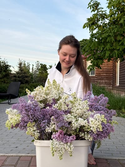 A woman is smiling and standing next to a white bucket filled with bunches of white and purple flowers. She is wearing a white jacket. The location is Ленино, Russia. In the background, there is a wooden house, a gray brick sidewalk, a tree, and a chair. The sky is visible.