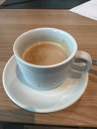 A white ceramic coffee cup, approximately two-thirds full of a light brown coffee beverage topped with a thin layer of crema, rests on a matching white ceramic saucer. Minor coffee staining is present on the interior rim and exterior surface of the cup, and a small brown speck is visible on the saucer. Both items are situated on a light-colored wooden table surface exhibiting a horizontal grain pattern. In the background, a dark, indistinct object and a partially visible white document with faint blue markings are discernible on the table. The scene is located in Kehl, Germany. No individuals or active interactions are depicted.