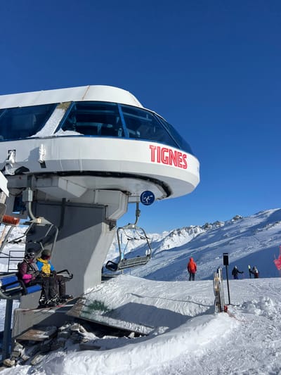 The image depicts a ski lift terminal at a ski resort in Tignes, France, on a clear, sunny day. The dominant feature is a large, white ski lift station, identified by the red "TIGNES" lettering on its front fascia, visible against a bright blue sky. A blue circular sign with a white icon of a skier is affixed to the terminal structure. Snow accumulates on portions of the terminal's blue-tinted panoramic windows.
Two individuals are seated on a blue and black chairlift positioned on the left side of the frame, waiting to depart or having just boarded. The individual on the left wears a pink or purple jacket, black pants, a black helmet, and goggles, with a snowboard attached to their feet. The individual to their right wears a yellow jacket, black pants, a black helmet, and goggles, with skis attached to their feet.
To the right of the lift station, on a wide, groomed, snow-covered slope, three additional individuals are visible. One person, clad in an orange jacket, stands facing away from the camera, holding ski poles, near what appears to be a queue entrance or exit point for the lift. Further right, two more individuals are partially visible, equipped with skis or snowboards. In the background, snow-covered mountains with visible rock formations extend under the clear blue sky. A vacant chairlift hangs further along the lift line, extending away from the terminal. The ground around the terminal and slope is entirely covered in undisturbed snow, with some shadows cast by the structures.