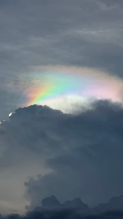 The image depicts a portion of the sky filled with dark, heavy clouds. A colorful, rainbow-like arc appears on the left side of the image. The location is given as Ba Ngoi Ward, Vietnam, but this information is not visually discernible from the image itself.