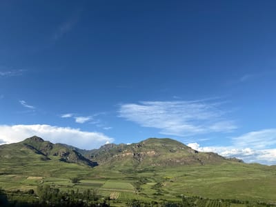 The image depicts a wide, sweeping landscape under a vast, clear blue sky with scattered white clouds. The foreground is dominated by rolling green hills and fields, with visible agricultural terraces and some trees along the bottom edge. In the middle ground, a prominent mountain range rises, characterized by steep, rocky slopes and lush green vegetation covering its lower sections. The mountains display distinct geological layers and formations. The overall scene is a natural, rural environment, suggesting a mountainous region. The provided crops confirm the detail of the mountain range and the expansive sky.