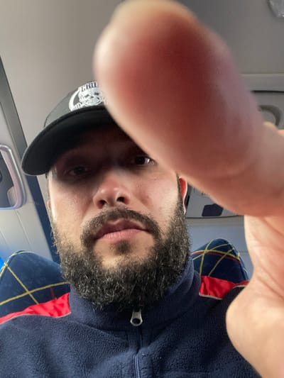 A man with a beard is wearing a blue jacket and a black baseball cap. He is in the interior of a vehicle, possibly a bus, as indicated by the patterned seats visible in the background. The man is holding up his hand, which is partially obscuring the camera lens, suggesting he is taking a selfie. The setting appears to be during the daytime, with natural light entering the vehicle. The image is cropped tightly on the man's face and upper body. There is no identifiable information to suggest the location is in Koskolovo, Russia.