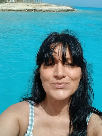 A woman with dark hair is visible against a turquoise background, likely water. She wears a light-colored garment with a checkered pattern. The setting appears to be in Sidi Abdel Rahman, Egypt, indicated by a glimpse of a shoreline and what might be infrastructure along the coast in the background.