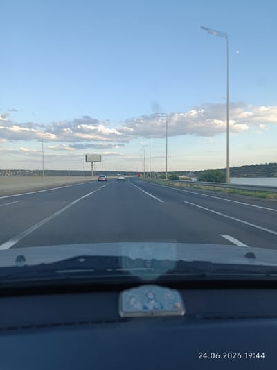This media file captures a view from the driver's seat of a vehicle traveling on a highway. The scene is a highway with multiple lanes, bordered by a concrete barrier on the left and a guardrail on the right, with a body of water visible in the distance on the right. The sky is a clear blue with scattered white clouds, and a crescent moon is faintly visible. Along the side of the highway, there are streetlights and a large billboard. Several cars are visible on the road, suggesting it is daytime and traffic is present.

A small religious icon is affixed to the dashboard in the foreground. The time and date stamp "24.06.2026 19:44" indicates the image was captured in the late evening on June 24, 2026. The general atmosphere is one of travel and movement. The location context suggests this highway is in or near Yelyzavetivka, Ukraine. The lighting indicates it is either late afternoon or early evening.