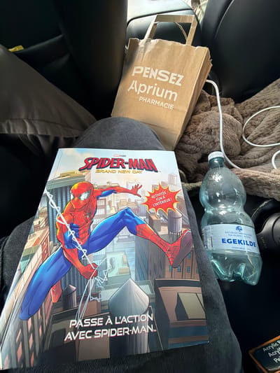 A close-up shot captures a person's lap in a car interior, with various items resting on it. The primary subject is a Spiderman comic book, featuring the character in a dynamic pose against a cityscape. To the right of the comic is a brown paper bag with white text that reads "PENSEZ Aprium PHARMACIE". Further to the right, a clear plastic bottle of EGEKILDE natural mineral water sits prominently, its label indicating it is from a source in the forest. A brown, textured blanket is draped behind the water bottle. The background shows the dark interior of a car, with the edge of a seat visible. The image appears to have been taken from a seated perspective within the vehicle, suggesting the person is either a passenger or the driver. The scene does not provide any specific visual cues to determine the location within the city of Ödsmål, Sweden, as there are no identifying landmarks or signs visible.