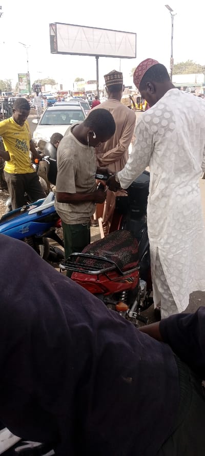 The image depicts an outdoor urban street scene in Fagge D2, Nigeria, occurring during daylight. In the foreground, the lower torso and legs of an individual wearing dark blue attire are partially visible.
The central activity involves four male subjects. A male wearing a light brown t-shirt, dark green trousers, and white in-ear headphones is bent forward, actively manipulating a black component located on the front section of a red motorcycle with both hands. The red motorcycle features a black and red patterned seat cover and a rear cargo rack.
Adjacent to the male working on the motorcycle, a second male, dressed in a white long-sleeved traditional garment with circular patterns and a red patterned cap, stands observing. His left hand is extended towards the area of activity on the motorcycle, and a silver-colored wristwatch is visible on his left wrist.
To the left of the central interaction, a third male in a yellow short-sleeved t-shirt bearing the text "JOMA YAMAHA BOYS" and dark trousers stands with hands on his hips, observing the scene. Further back, a fourth male in a light brown traditional garment and a patterned cap is visible, facing away from the viewer.
The background reveals a moderately busy street environment with vehicular traffic, including a white sedan and a yellow auto-rickshaw (Keke Napep), along with several distant pedestrians. A large, blank rectangular billboard with a black frame is prominently positioned in the upper background. To its left, a smaller green billboard depicting two individuals is visible. A blue and white motorcycle is partially seen on the left side of the frame, behind the man in the yellow shirt. A street light pole with a solar panel is present on the right side of the background. The ground appears to be a paved surface.