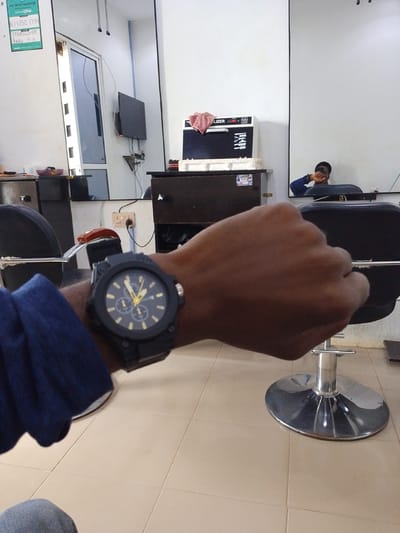 The image depicts the interior of a barbershop in Abeere, Nigeria. The focus is on a person's wrist wearing a black, chunky wristwatch with a black face and yellow accents. The watch has three subdials. In the background, reflected in a large mirror, a person is seen sitting in a barber's chair, taking a selfie with their phone. The barbershop environment is visible, including barber chairs, cabinetry, and a wall-mounted television. The floor is tiled.