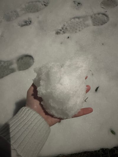 In an outdoor setting within the city of Visp, Switzerland, a human left hand, clad with a light-colored, ribbed sweater cuff, holds a significant, irregular clump of fresh, crystalline snow. The snow-covered ground around the hand displays multiple distinct footprints from at least one individual, with visible sole patterns impressed into the white surface. Small, dark organic debris is sparsely embedded in the snow. The scene is illuminated by an artificial light source, casting a pronounced shadow from the hand onto the snow below and causing specular reflections on the snow crystals.