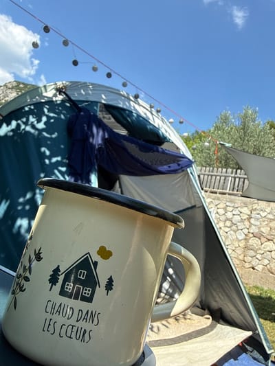 In the city of Kanjoši, Montenegro, a close-up shot features an enamel mug in the foreground, displaying a drawing of a house, trees, and the French phrase "CHAUD DANS LES CŒURS." Behind the mug, a tent is visible, with a blue fabric draped inside. String lights with round bulbs hang across the sky. The background includes a stone wall, greenery, and a clear blue sky with scattered clouds. The scene appears to be outdoors, possibly at a campsite.