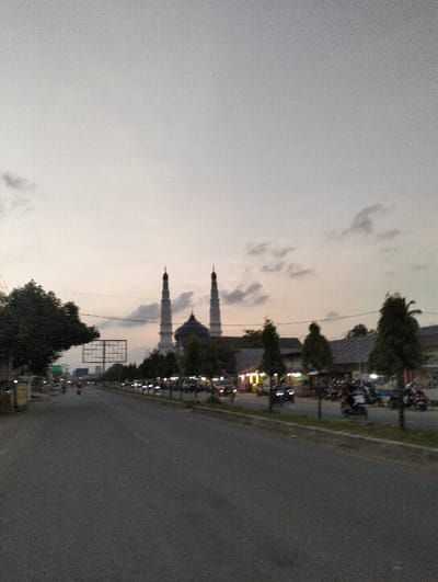 The image displays an urban street scene in Payacut, Indonesia, observed during dusk or dawn. A wide, dark asphalt road dominates the foreground and extends into the mid-distance. In the background, a large mosque featuring a central dome and two prominent minarets is visible, appearing light-colored against the fading sky. The sky is largely overcast, showing a gradient from light grey to pale orange near the horizon with scattered clouds.

Along the right side of the road, a planted median contains a uniform row of conical trees. Behind these trees, multiple single and two-story commercial buildings are present, some exhibiting artificial illumination. A collection of motorcycles and a few cars are either parked or in motion on this side of the street. On the left side of the road, a large, empty billboard structure is discernible in the distance, alongside sporadic foliage. A single motorcycle with a rider is observed in transit in the left lane, heading away from the viewer. Additional vehicles, including cars with activated headlights, are visible further down the road, indicating ongoing traffic.