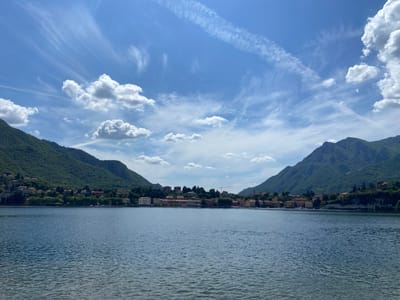 The image depicts a serene lake scene near the city of Lecco, Italy. The foreground is dominated by a large expanse of water with subtle ripples on its surface. In the midground, a line of buildings sits along the shoreline, possibly a town or village on the lake's edge. The buildings appear to be a mix of residential and possibly commercial structures with red-tiled roofs. The background features two prominent, densely forested mountains that frame the lake, adding depth and dimension to the scene. The sky is blue with a mix of puffy cumulus clouds and faint contrails, suggesting a clear day with air traffic. There are no people visible in the image. The overall environment suggests a calm and picturesque setting.