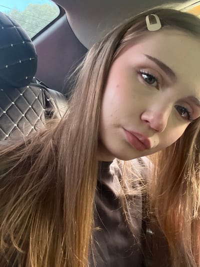 A close-up, eye-level, medium shot captures a young woman with light brown hair, styled with a light-colored barrette, looking directly at the camera. Her face is lightly made up with blush and eyeliner. She is seated in the interior of a vehicle, as indicated by the quilted headrest and window frame visible on the left side of the frame. The background outside the window appears to be foliage and sky, suggesting an outdoor setting. The lighting is somewhat dim, possibly indicating it is daytime. The specific location in Novaya Otradovka, Russia, cannot be determined from the visual information provided.