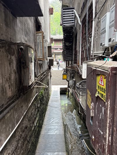 The image depicts a narrow alleyway in Fenghuang, China. The buildings flanking the alley are densely packed with electrical wiring, utility boxes, and air conditioning units. A narrow concrete pathway runs through the alley, with a shallow stream or runoff alongside it. The walls adjacent to the stream are covered in moss and vegetation. Stairs are visible at the end of the alleyway, leading upward. Trees and other greenery are also visible. A warning sign in Chinese characters is attached to one of the buildings. There are some figures on the stairs in the distance.