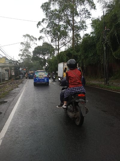 The image displays a traffic scene on a wet asphalt road in the city of YonPhoogla, Bhutan, under an overcast sky. In the immediate foreground, a person is seen from the rear, riding as a passenger on a dark-colored scooter. The passenger wears a black helmet, a red long-sleeved jacket, and dark blue and white plaid pants, with white sneakers visible on their feet. The road surface is dark and reflective from wetness, featuring a continuous white line along its left edge.
Ahead of the scooter, a blue hatchback automobile, adorned with various decals on its rear window, proceeds in the same direction. Further along the road, a light-colored box truck and several other motorcycles are visible in the flow of traffic. The left side of the road features several low-rise structures, some appearing weathered, with a network of overhead utility wires. The right side of the road is demarcated by a dense embankment of green foliage, comprising tall trees and various vines, alongside a dark utility pole.