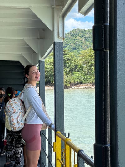 A young Caucasian woman with dark hair pulled back in a ponytail smiles at the camera while standing on a ferry in Thailand. She is wearing a white long-sleeved shirt, pink athletic shorts, and a backpack with a cartoon print. Behind her, other passengers are visible. The ferry is traveling on calm, light blue water. In the distance, a lush green, mountainous island is visible, with trees growing down to the water's edge. The sky is partly cloudy with white clouds. The foreground of the image shows the railing and structural elements of the ferry, including black metal poles and a yellow railing. The overall scene suggests a journey through a tropical landscape.