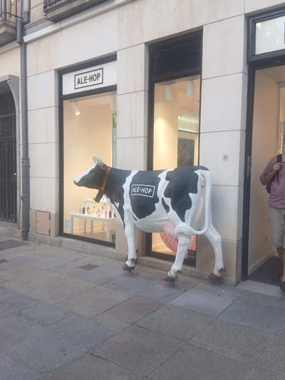 A life-sized, black and white cow statue is positioned on a sidewalk in front of a storefront. The cow has a black collar and appears to be chained by its hind legs to the ground. A person, wearing a pink shirt and tan shorts, stands in the background, partially visible. The storefront has large windows displaying items inside. The signage visible on the storefront reads "ALE-HOP". The sidewalk is paved with stone tiles. The lighting suggests it is daytime. The scene appears to be a commercial area in Ávila, Spain.