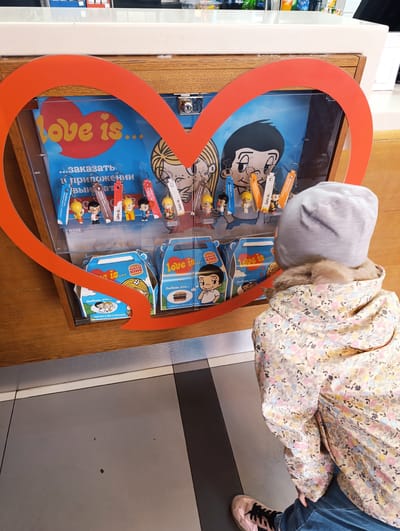 Here is a forensic description of the image:

The image shows a young child, viewed from the back, standing in front of a display case decorated with a large red heart. The display contains "Love is..." themed merchandise, including keychains and Burger King kids' meal boxes, inside of what appears to be a Burger King restaurant. The merchandise is decorated with the classic "Love is..." cartoon characters. 

The child is wearing a light grey beanie, a floral-patterned jacket with a fur-trimmed hood, and blue pants. They are also wearing pink sneakers with black laces. The display case has the "Love is..." logo and text in Russian. 

The floor is tiled in grey and black and in the background are details of the restaurant's service counter. The image was taken in the city of Noginsk, Russia.