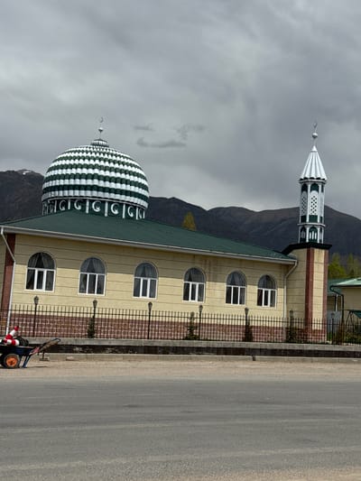 The image shows a light-colored mosque with a large, green-and-white striped dome and a smaller minaret with a white spire. The mosque has a green roof and arched windows. A black, wrought-iron fence runs along the side of the mosque, separating it from a gray asphalt road. A cart with orange wheels and red-and-white striped poles is parked near the fence. In the background, there are mountains under a cloudy gray sky. The location is the city of Temir, Kyrgyzstan.
