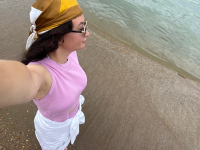 A woman stands on a sandy shore next to calm, shallow water. She is facing right, looking out at the water. She has dark curly hair, which is partially covered by a patterned headscarf with shades of brown, yellow, and white. She wears large, round, gold-rimmed sunglasses with black arms that say "TOM FORD." Her top is a bright pink, knitted, sleeveless shirt. Around her waist, she has a white cover-up or sarong. Her left arm is extended, holding a phone to take a selfie. The background consists of the sandy beach and the light blue-green water, with a slight ripple on the surface. The overall lighting is overcast. The location is a beach, potentially in Derbent, Russia, given the context.