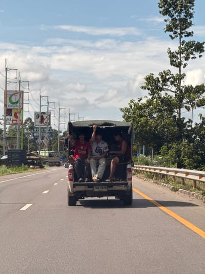 In Tambon Tako, Thailand, four young males are seen riding in the open bed of a white pickup truck on a paved road. The truck is moving forward, indicated by motion blur in the background and the road markings. The sky is partly cloudy with a blue hue. On the left side of the road, there are overhead power lines and various signs, including a prominent "bangchak" sign and others with Thai script. A tall tree with dense foliage stands on the right side of the road, bordering a yellow and white guardrail.

The individuals in the truck are dressed casually. The two in the front are wearing a red soccer jersey with "Liverpool" and "Standard Chartered" printed on it, and a white t-shirt with the number "66" on it. The person in the red jersey is looking towards the front of the truck, while the person in the white shirt is looking to the right. The two individuals behind them are less clearly visible. One appears to be wearing a dark sleeveless shirt and shorts, and the other is wearing a dark t-shirt and shorts. The truck has a dark canvas cover over the rear. The license plate of the truck is visible and reads "บข 7879". The road has white lane markings and a solid yellow line on the right edge. The environment suggests a rural or semi-urban setting with both natural elements and infrastructure like power lines and signs.