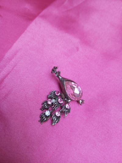 A silver-toned metal brooch, depicting a peacock, is positioned centrally on a bright pink textile surface. The peacock's body is composed of a large, faceted, teardrop-shaped pink gemstone. Its tail features five distinct feather-like elements, each set with a smaller, round, faceted gemstone; four are pink, and one is clear or very light blue. The metal components exhibit an antiqued finish. The underlying pink fabric is plain and shows soft folds, filling the entire frame. The scene is located in Qarabucaq qəsəbə, Azerbaijan.