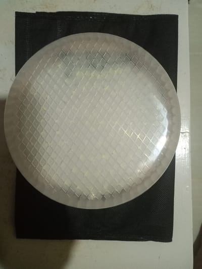 The image displays a circular, translucent object with a raised diamond-grid pattern, possibly a diffuser cover for an LED light fixture. Through the translucent material, a grid of numerous small, yellowish-white rectangular components, consistent with individual LED emitters, is visible. This circular object is placed upon a rectangular piece of black, textured, non-woven fabric. The black fabric rests on a light-colored surface, which transitions from a mottled light gray on the upper left to a smoother, off-white surface on the right and bottom, suggesting an adjacent wall or countertop. No people are present, and no specific actions or interactions are occurring. The image does not contain any visual information to determine its geographical location in Uleeceubrek, Indonesia, or anywhere else.